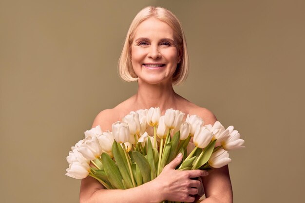 Foto portret van een mooie blonde vrouw van middelbare leeftijd die een boeket witte tulpen vasthoudt en glimlacht op een olijfachtergrond