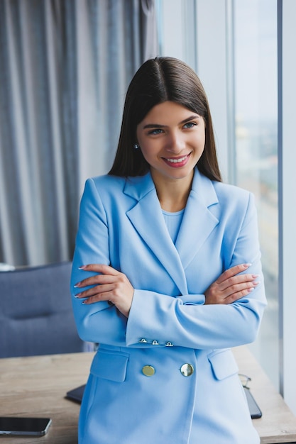 Portret van een mooie brunette in een blauwe jas op kantoor in de buurt van het panoramische raam met uitzicht op de stad Gelukkige zakenvrouw op kantoor