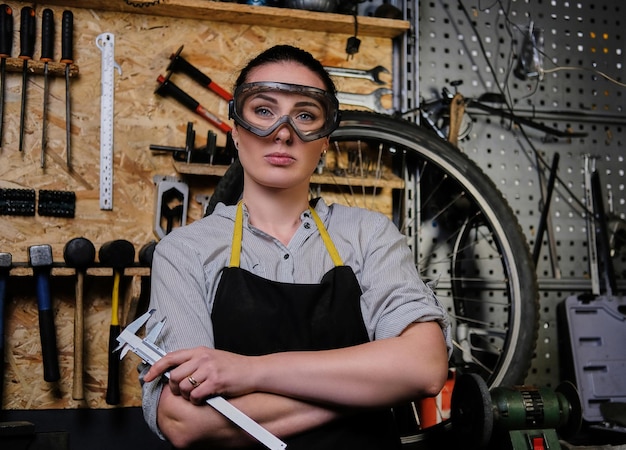 Portret van een mooie brunette vrouw die werkkleding, schort en bril draagt, houdt een remklauw vast en staat in een werkplaats tegen muurgereedschap.