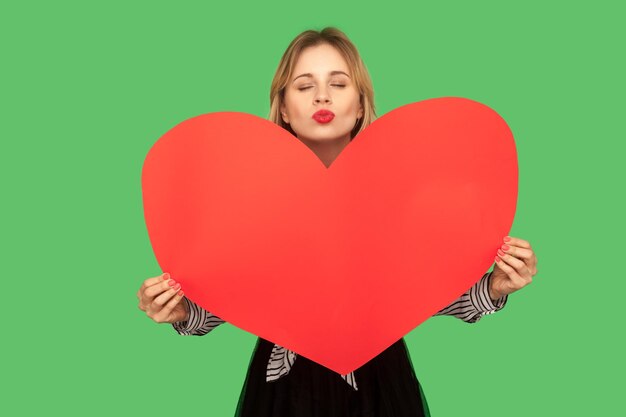 Portret van een mooie charmante vrouw die een groot rood hart vasthoudt en een luchtkus stuurt met sexy rode lippen, liefde deelt, amoureuze gevoelens op Valentijnsdag. indoor studio-opname geïsoleerd op groene achtergrond
