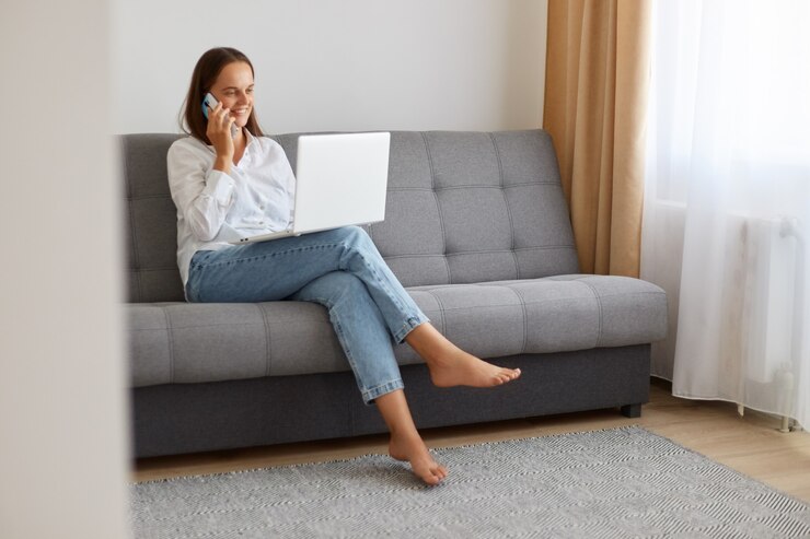 Foto portret van een mooie donkerharige vrouw die op de mobiele telefoon spreekt voor haar laptopcomputer thuis terwijl ze op de bank zit in een lichte woonkamer, met een positieve gezichtsuitdrukking.