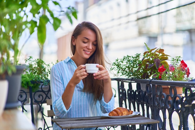 Portret van een mooie gelukkig schattige vrolijke lachende romantische vrouw met een aromatische koffiekopje in handen en een bord met vers gebakken croissant op de tafel in de ochtend op een balkon