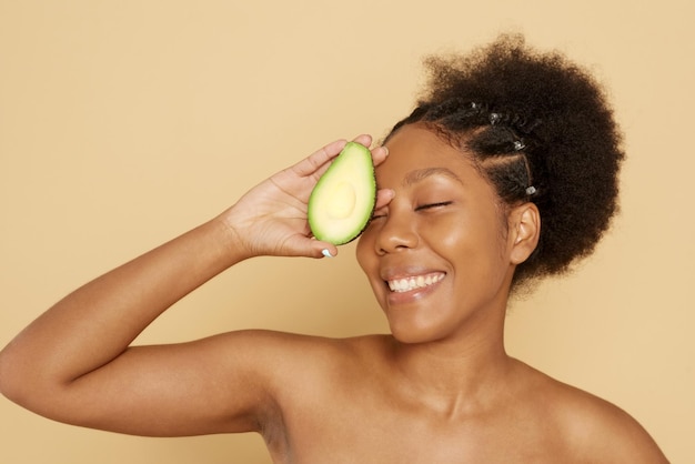 Portret van een mooie jonge Afrikaanse vrouw die een avocado vasthoudt en lacht tegen een beige achtergrond Het concept van de juiste voeding, huidverzorging, spa en cosmetologie