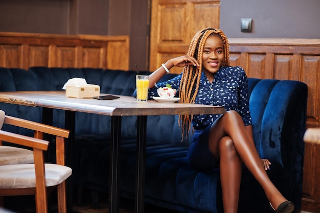 Portret van een mooie jonge afrikaanse zakenvrouw met dreadlocks die een blauwe blouse en rok draagt die in café zit met ijs en ananassap