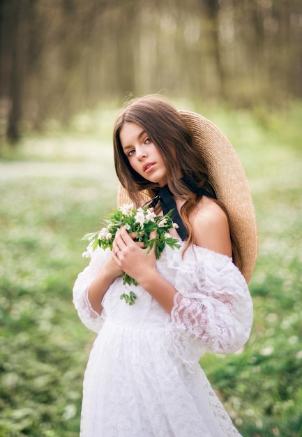 Portret van een mooie jonge brunette in een lentebos Een meisje met een boeket sleutelbloemen in haar handen in een witte kanten jurk