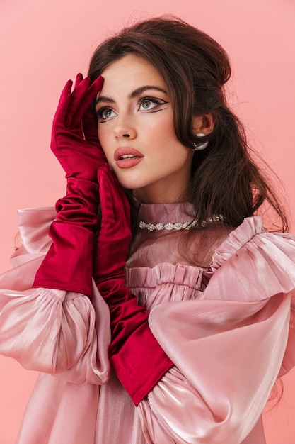 Portret van een mooie jonge brunette vrouw met lichte make-up die modieuze kleding draagt, geïsoleerd over roze muur, poserend