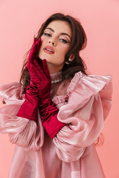 Portret van een mooie jonge brunette vrouw met lichte make-up die modieuze kleding draagt, geïsoleerd over roze muur, poserend