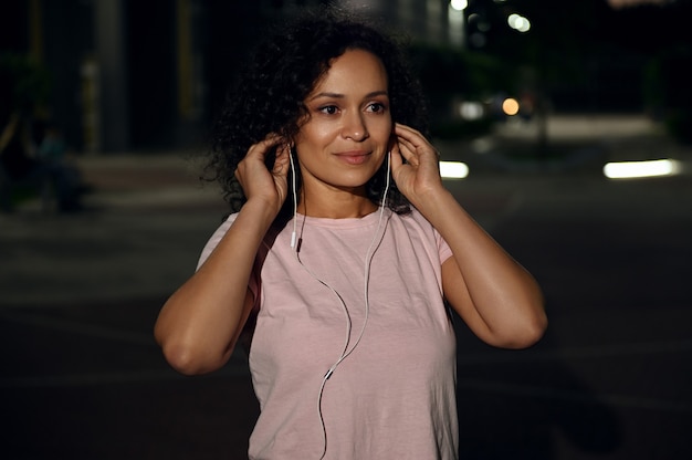 Portret van een mooie jonge Latijns-Amerikaanse vrouw die oortelefoons draagt en wegkijkt, staande op de achtergrond van de avondverlichting