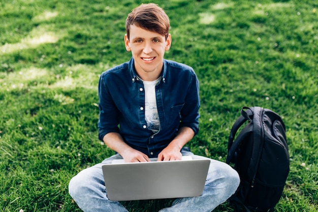 Portret van een mooie jonge man zittend op het gras in een park met een laptop