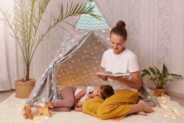 Portret van een mooie jonge volwassen moeder die een boek vasthoudt en een sprookje leest voor haar dochtertje dat op de vloer ligt en probeert in slaap te vallen familie die de avond samen doorbrengt