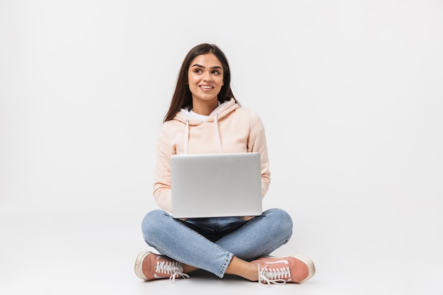 Portret van een mooie jonge vrouw casual gekleed zittend, benen gekruist geïsoleerd op wit, met behulp van laptop computer