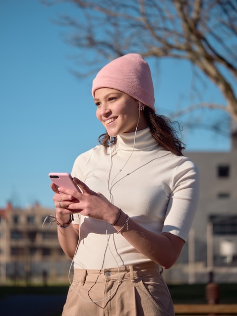 Portret van een mooie jonge vrouw die een roze hoed en vrijetijdskleding draagt, een mobiele telefoon met oortelefoons gebruikt en er gelukkig en ontspannen uitziet