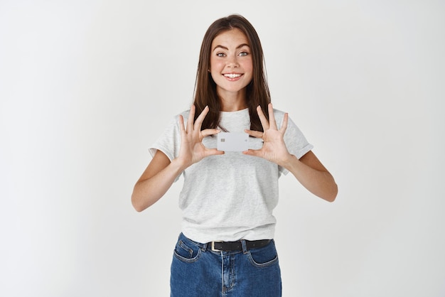 Portret van een mooie jonge vrouw gekleed in een t-shirt en spijkerbroek met creditcard en glimlachend in de camera witte achtergrond