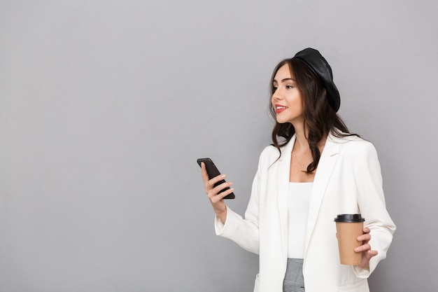 Portret van een mooie jonge vrouw gekleed in jas over grijze achtergrond, met kopje koffie, met behulp van mobiele telefoon, wegkijken