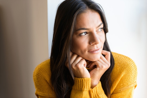 Portret van een mooie jonge vrouw gekleed in trui zit bij het raam binnenshuis close-up