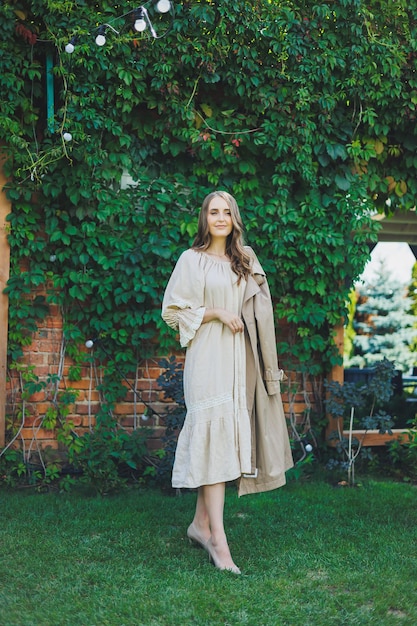 Portret van een mooie jonge vrouw in een lichte beige jurk Romantische vrouw in een jurk met een glimlach op haar gezicht op de achtergrond van een groene tuin Vrouwelijke cocktailjurk voor een vakantie