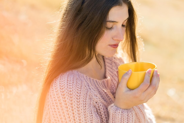 Portret van een mooie jonge vrouw in een stijlvolle gebreide trui die op een zonnige dag op het gouden herfstbos staat. tijd om te ontspannen en wat koffie te drinken