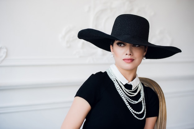 Portret van een mooie jonge vrouw in retro stijl in een elegante zwarte hoed en jurk over luxe rococo muur