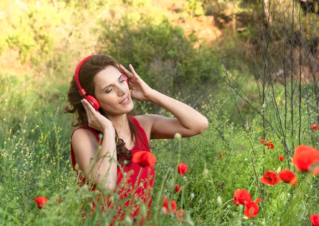 Portret van een mooie jonge vrouw met gesloten ogen, zittend in een veld met klaprozen, luisterend naar muziek op haar koptelefoon