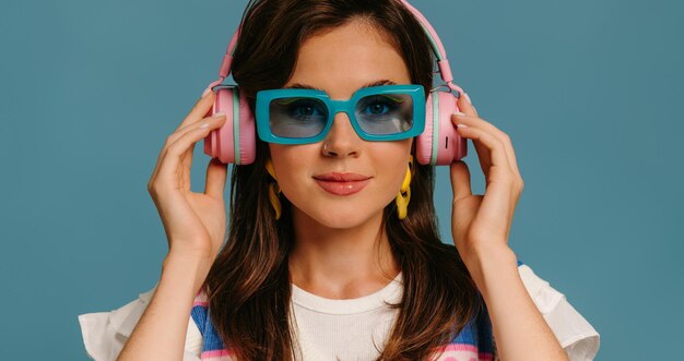 Portret van een mooie jonge vrouw met koptelefoon en trendy bril die van muziek geniet op een blauwe achtergrond