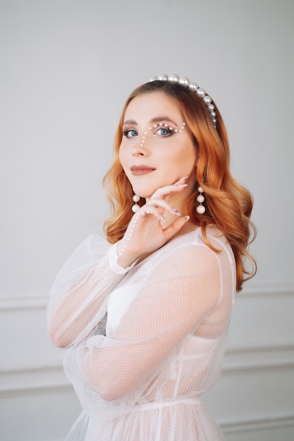 Portret van een mooie jonge vrouw met rood haar in een witte jurk en make-up gemaakt van parels op haar ...