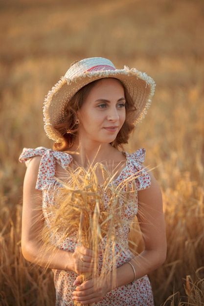 Portret van een mooie jonge vrouw Vrouw in jurk en hoed poseren in tarweveld bij zonsondergang