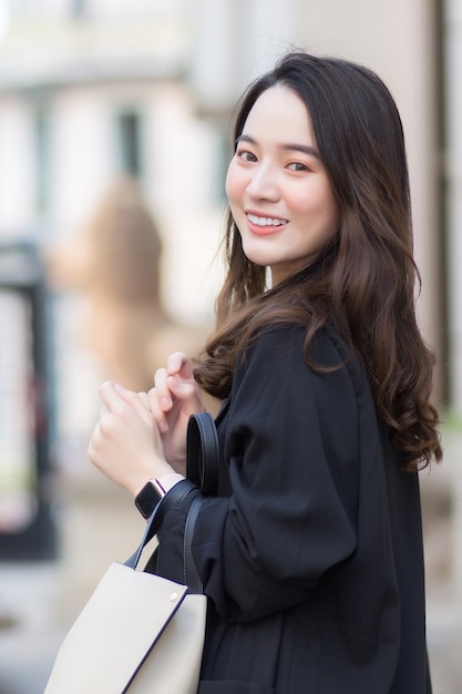 Portret van een mooie langharige Aziatische vrouw in een zwarte jas met een wandeltas glimlachend