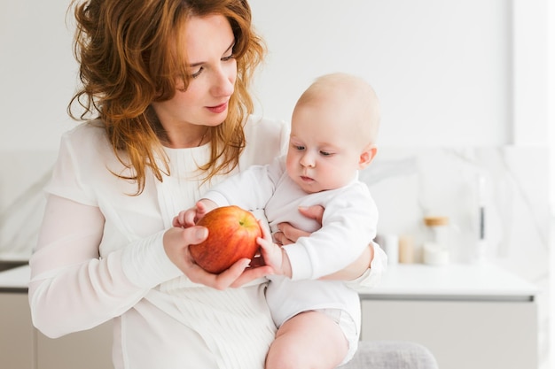 Portret van een mooie moeder die op de keuken staat en haar schattige kleine baby en grote rode appel in handen houdt