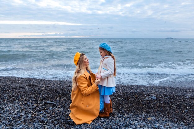 Portret van een mooie moeder en dochter in warme kleren aan de kust in het koude seizoen, familiewandeling langs het strand