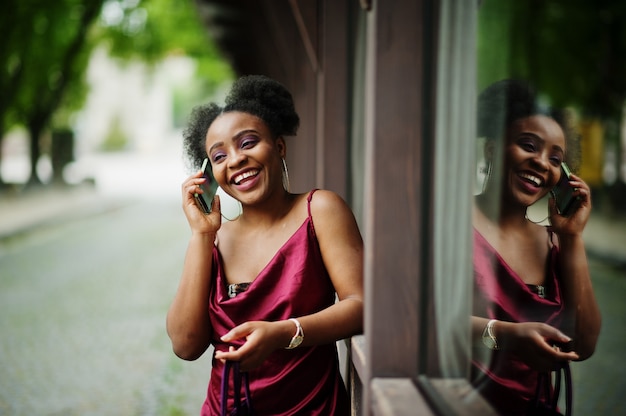 Portret van een mooie natuurlijke jonge afrikaanse vrouw met afrohaar. zwart model in rode zijden jurk met mobiele telefoon.