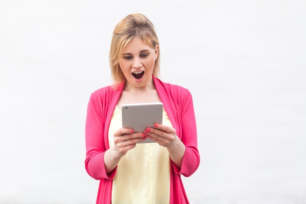 Portret van een mooie ongelooflijke jonge vrouw in een roze blouse die staat en kijkt naar verraste video op haar tablet met een geschokt gezicht. Binnen, geïsoleerd, studio-opname, witte achtergrond