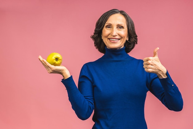 Portret van een mooie oudere volwassen leeftijd vrouw met een appel, glimlachend, geïsoleerd over roze achtergrond. Een appel per dag houdt een dokter weg.