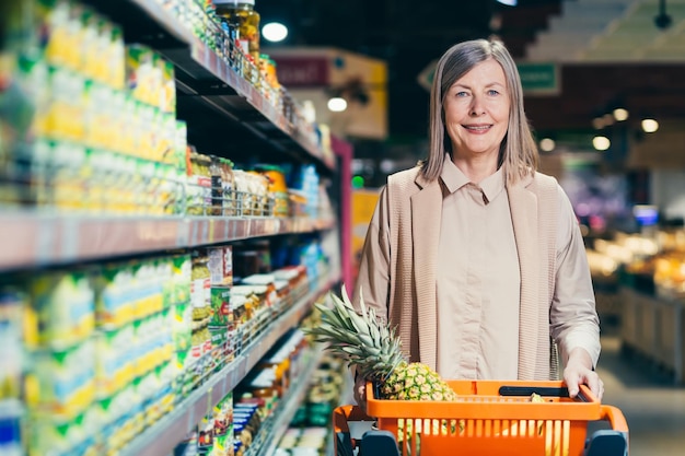 Portret van een mooie oudere vrouw die tussen de schappen van een winkelsupermarkt staat met een mand vol voedselfruit Hij kijkt naar de camera en glimlacht