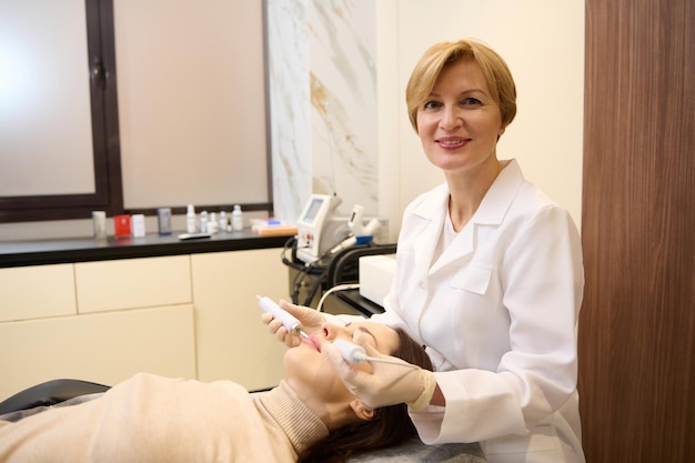 Portret van een mooie rijpe blonde vrouw arts schoonheidsspecialist doen anti-aging huidverzorging behandeling aan vrouw gezicht met behulp van elektrische impulsen microstroom behandeling hardware cosmetologie concept