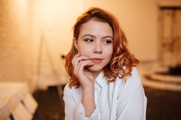 Portret van een mooie roodharige jonge vrouw gekleed in een wit overhemd opzij kijkend met de hand die haar gezicht aanraakt