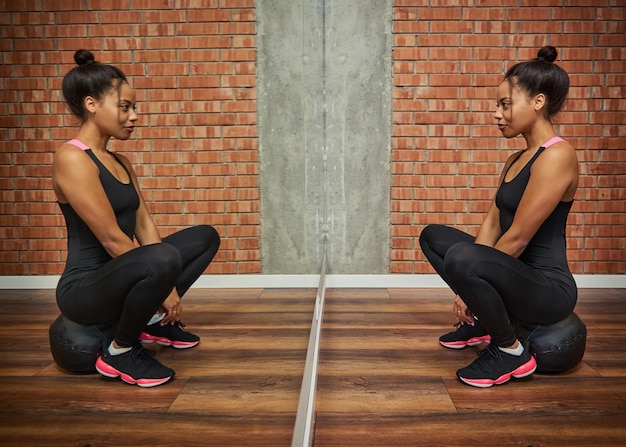 Portret van een mooie sportieve atleet Afrikaanse vrouw in zwarte sportkleding kijkend naar haar spiegelreflectie, zittend op een medicijn fitness bal tegen rode bakstenen muur achtergrond van een gymles