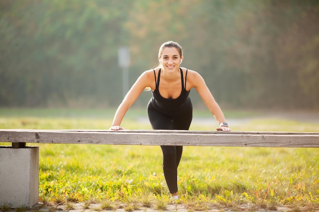 Portret van een mooie sportieve vrouw van 20 jaar in sportkleding die push-ups doet en naar muziek luistert met bluetooth-oordopjes tijdens de training in het groene park.