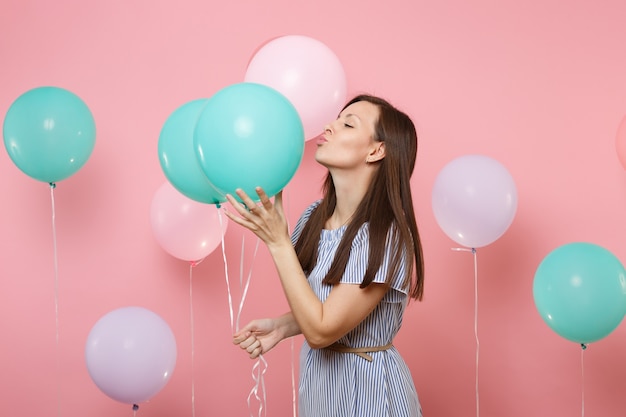 Portret van een mooie tedere brunette vrouw, gekleed in een blauwe gestreepte jurk, zoenend met kleurrijke luchtballonnen op een heldere trending roze achtergrond. Verjaardagsfeestje, mensen oprechte emoties concept.