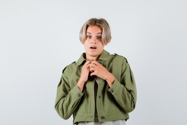 Portret van een mooie tienerjongen die zijn handen op de borst houdt in een groene jas en er bang uitziet vooraanzicht