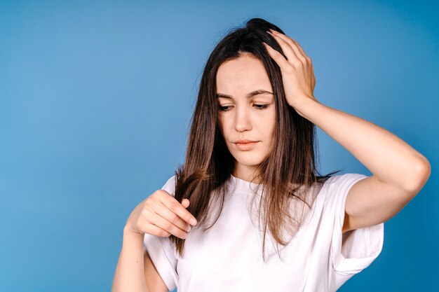 Portret van een mooie trieste jonge vrouw met lang haar in haar hand. Close-up Ongelukkig vrouwelijk model kijken naar gespleten haar. Haar zorg concept.