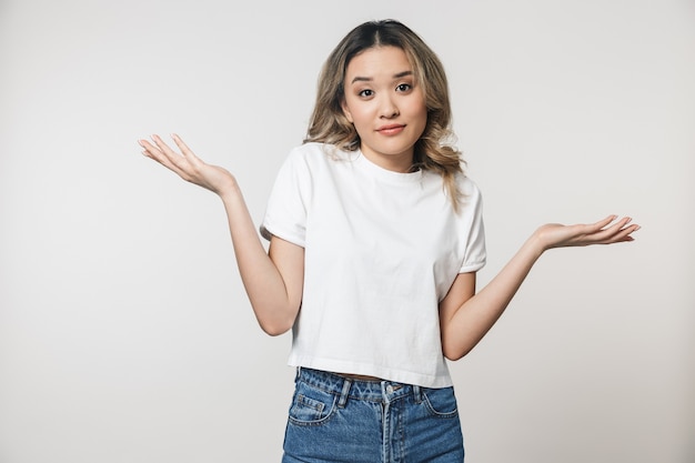 Portret van een mooie verwarde jonge aziatische vrouw die geïsoleerd over een witte muur staat, schouders ophalend