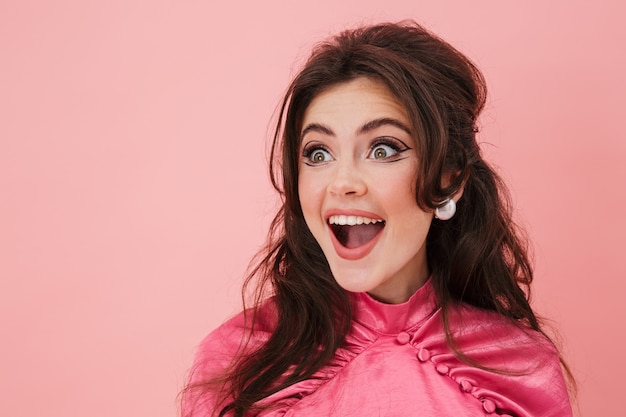 Portret van een mooie, vrolijke jonge brunette vrouw met lichte make-up die modieuze kleding draagt, geïsoleerd over een roze muur, wegkijkend