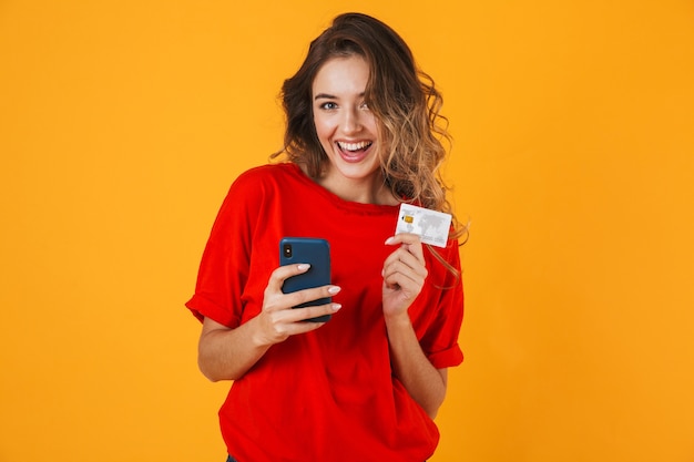 Portret van een mooie vrolijke jonge vrouw die geïsoleerd over een gele muur staat en een creditcard toont tijdens het gebruik van een mobiele telefoon