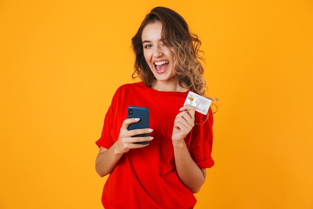 Portret van een mooie vrolijke jonge vrouw die geïsoleerd over een gele muur staat en een creditcard toont tijdens het gebruik van een mobiele telefoon