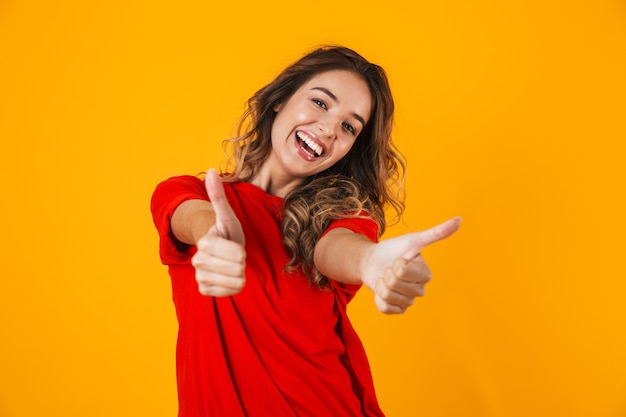 Portret van een mooie vrolijke jonge vrouw die geïsoleerd staat over een gele muur, duimen omhoog