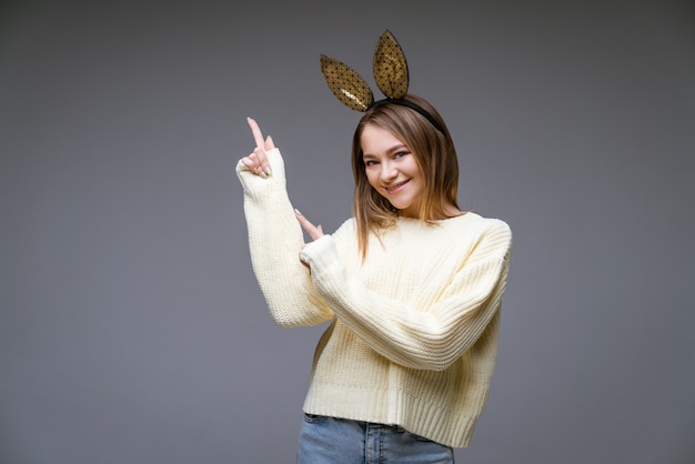 Portret van een mooie vrolijke jonge vrouw in een trui en in konijnenoren toont haar vinger opzij op een grijze scène