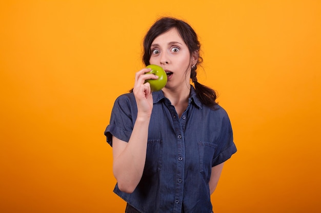 Portret van een mooie vrouw die een groene appel bijt en naar de camera in de studio kijkt over gele achtergrond. jonge vrouw die gezond fruit eet. vrolijke vrouw die een organiserende appel eet.