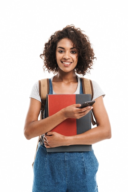 Portret van een mooie vrouw die een rugzak draagt met oefenboeken en een mobiele telefoon die over een witte muur wordt geïsoleerd