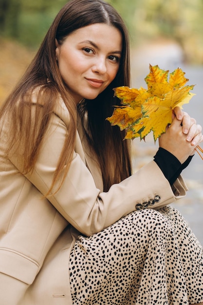 Portret van een mooie vrouw in een beige jas die gele esdoornbladeren vasthoudt terwijl ze in een herfstpark zit
