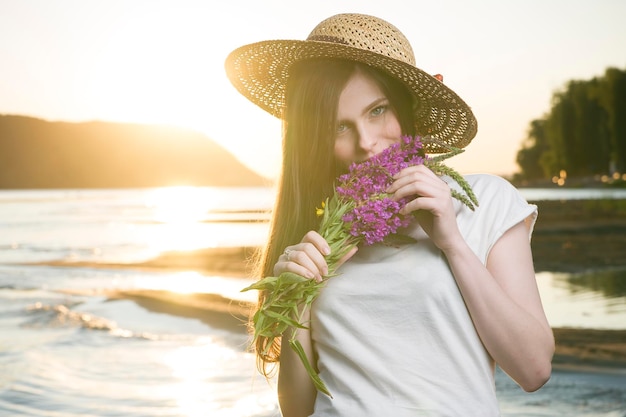Portret van een mooie vrouw in een hoed op een achtergrond van een zonsondergang. Meisje met een boeket bloemen